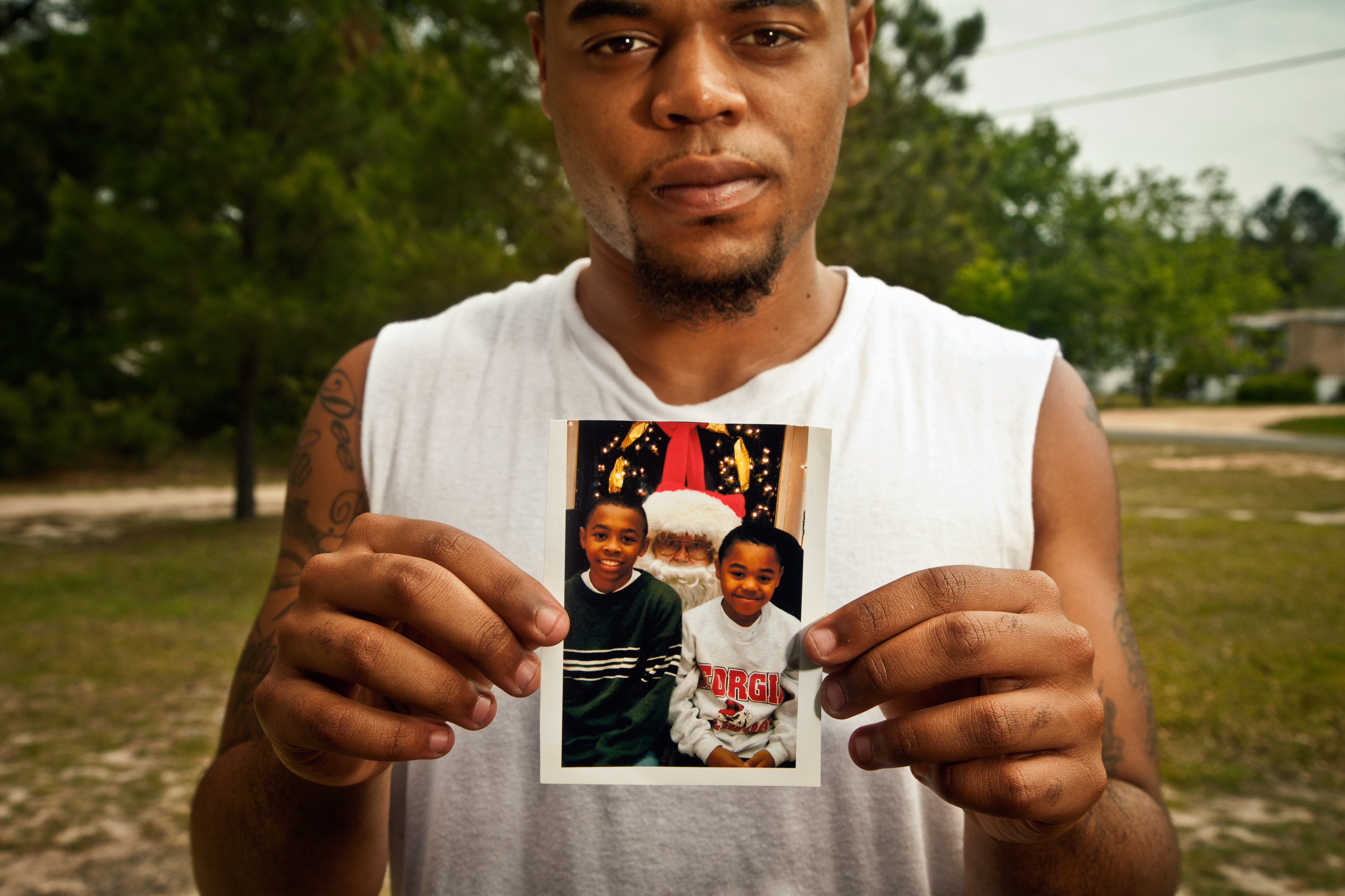 A man in a white tank top holding up a photograph of himself and his brother