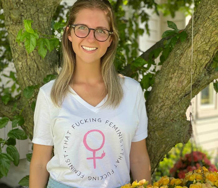 A woman in glasses smiles while wearing a shirt that reads "That fucking feminist."