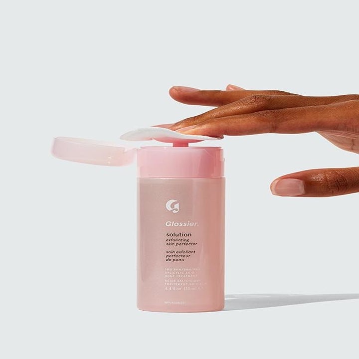 On the right, a hand presses a makeup remover bad on top of a pink bottle that says "Glossier Solution Exfoliating Skin Perfector" 