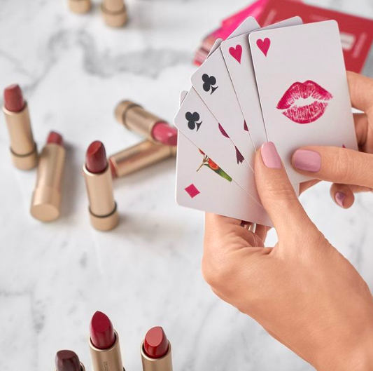 Several tubes of lipstick out on a table while a person holds playing cards above them 
