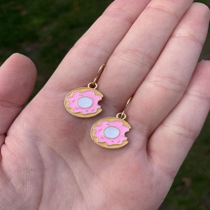 hand holding two earrings shaped like a donut with pink icing on top and a bite taken out of them
