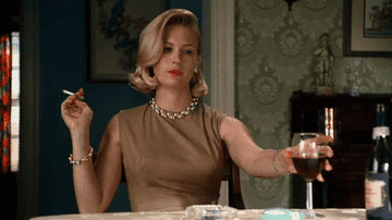 Betty Draper sitting at a table picking up a glass of red wine while smoking a cigarette. 