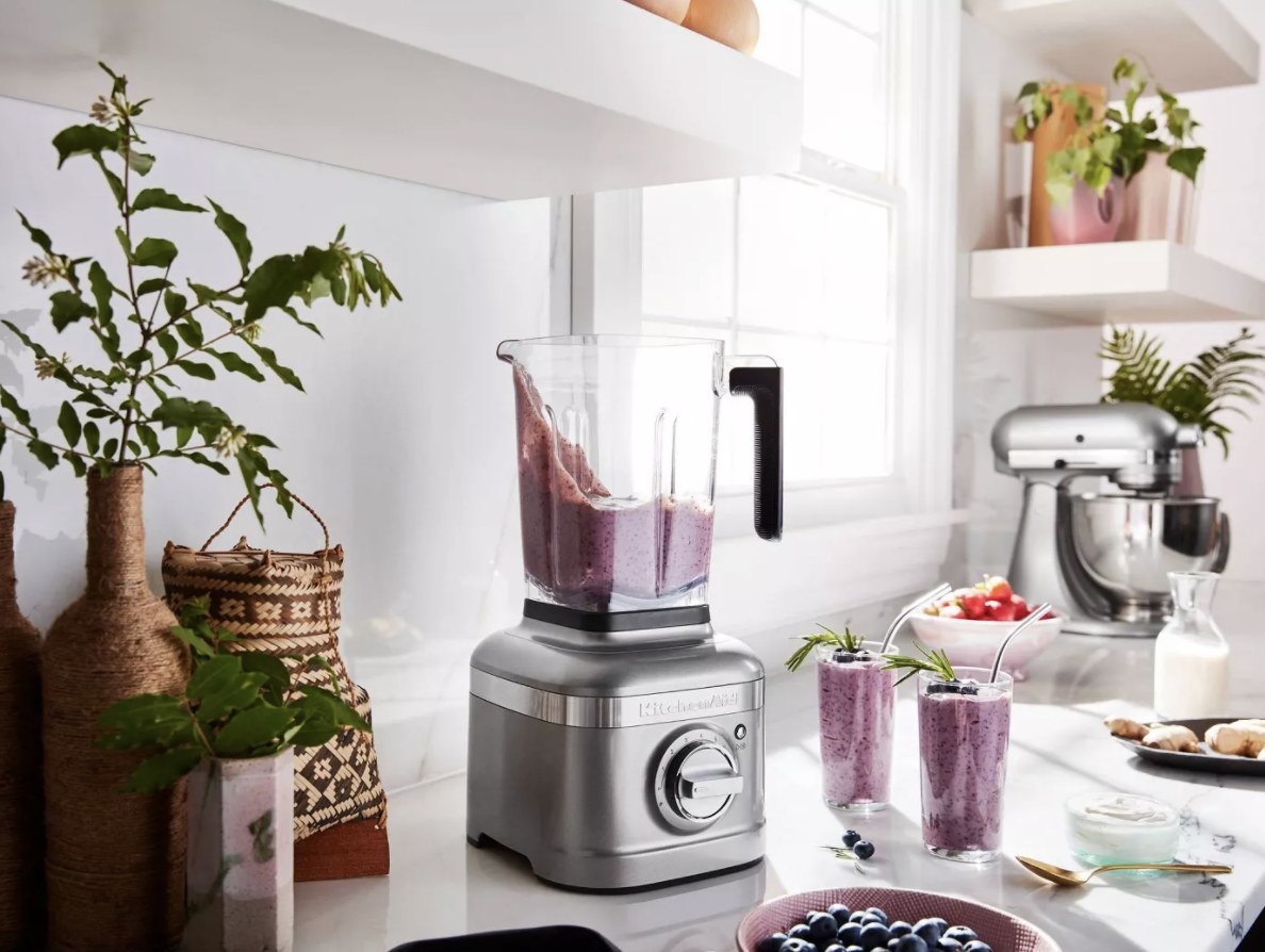 the blender with the ingredients for a blueberry smoothie laid our in front of it,  blueberry smoothie mix inside of it, and fresh glasses of blueberry smoothies next to it 