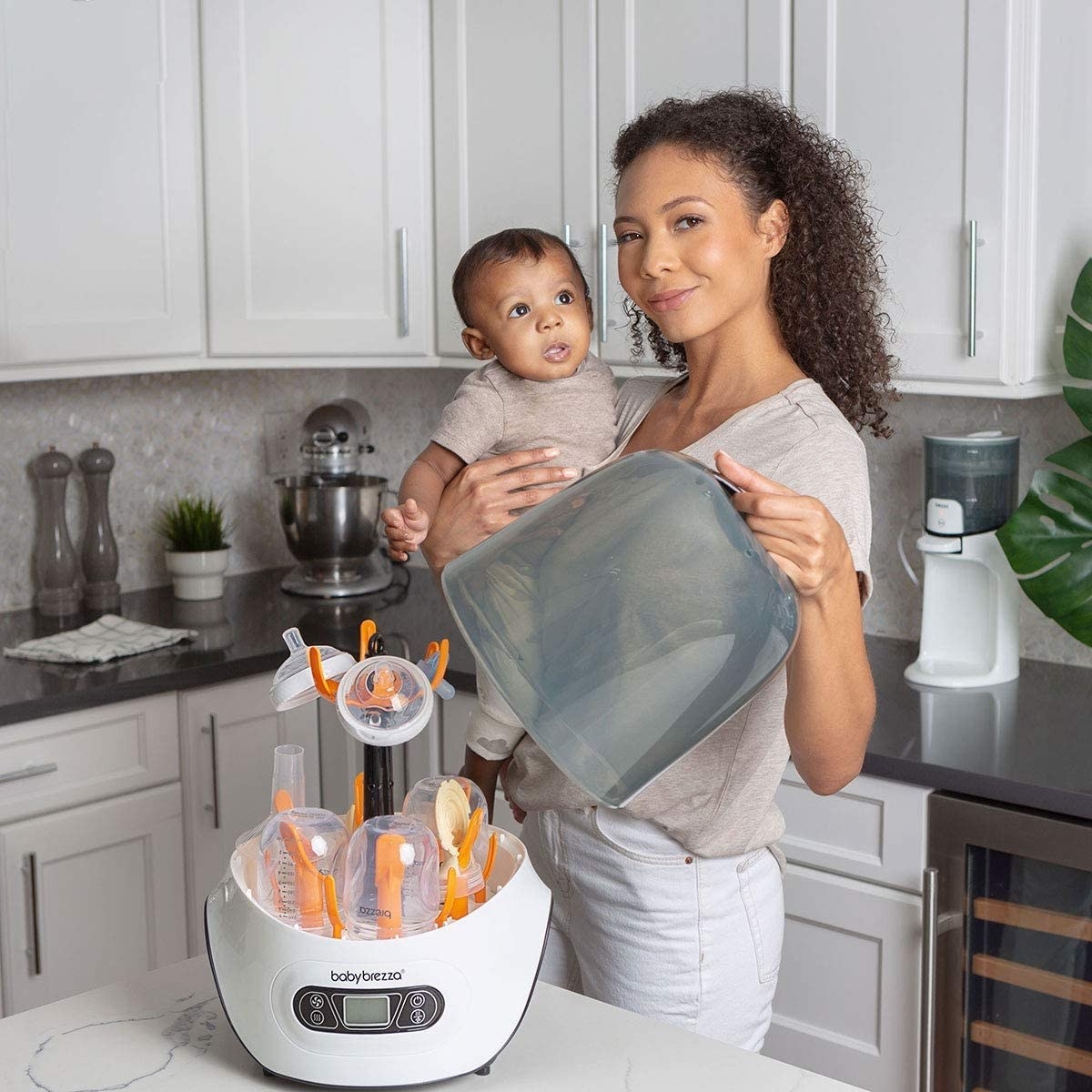 A person holding a baby and lifting the lid off an electric bottle sterilizer