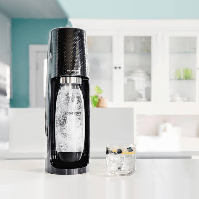 The black machine with a water bottle inside and bubbles, showing that it&#x27;s carbonating the water with a glass of sparkling water with fruit next to it