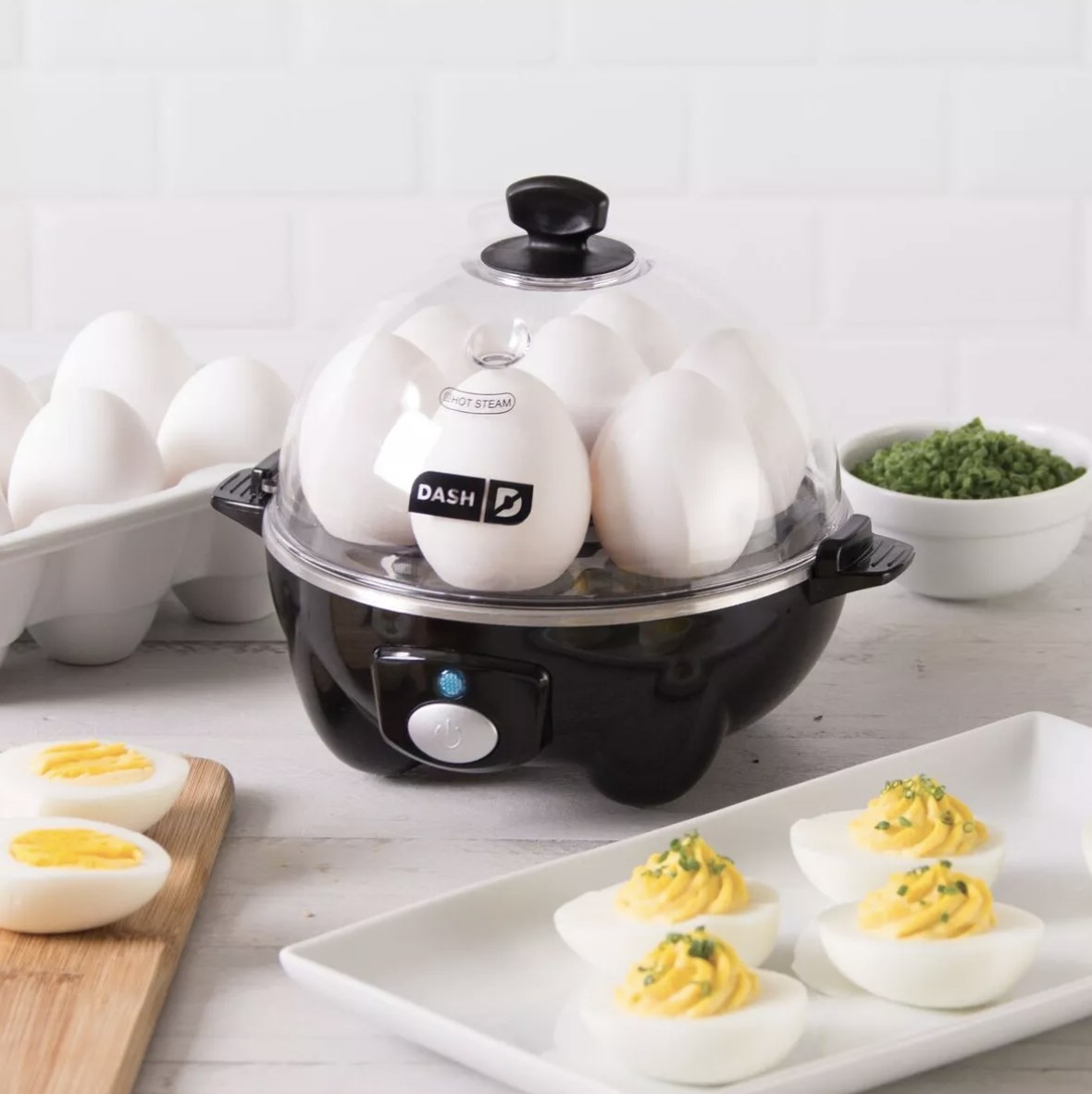 the egg cooker with eggs in a carton behind it, eggs inside of it cooking, and deviled eggs on a plate next to it 
