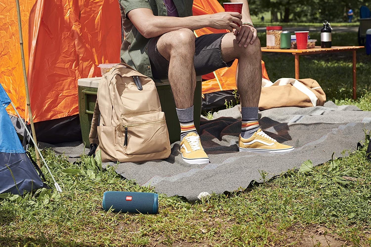 A person sitting on a cooler with a portable speaker on the grass in front of them