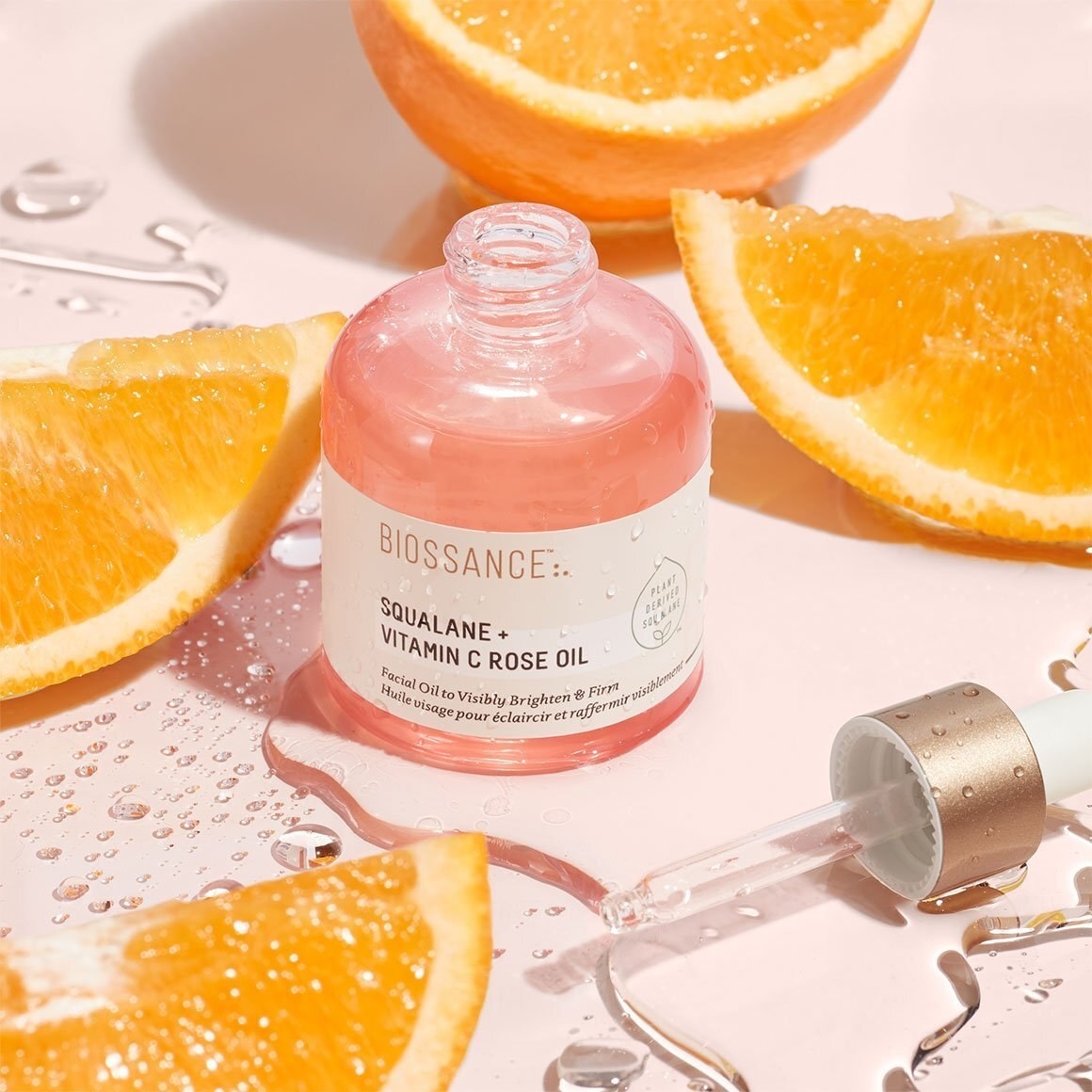 Pink dropper bottle of Biossance Squalane + Vitamin C Rose Oil next to orange slices