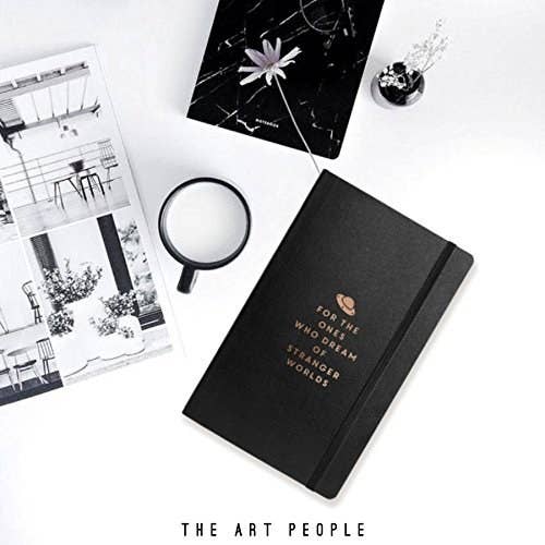 A black journal which has the following phrase written in gold on its cover, "For the ones who dream of stranger worlds". It's pictured with a mug, poster and plant.
