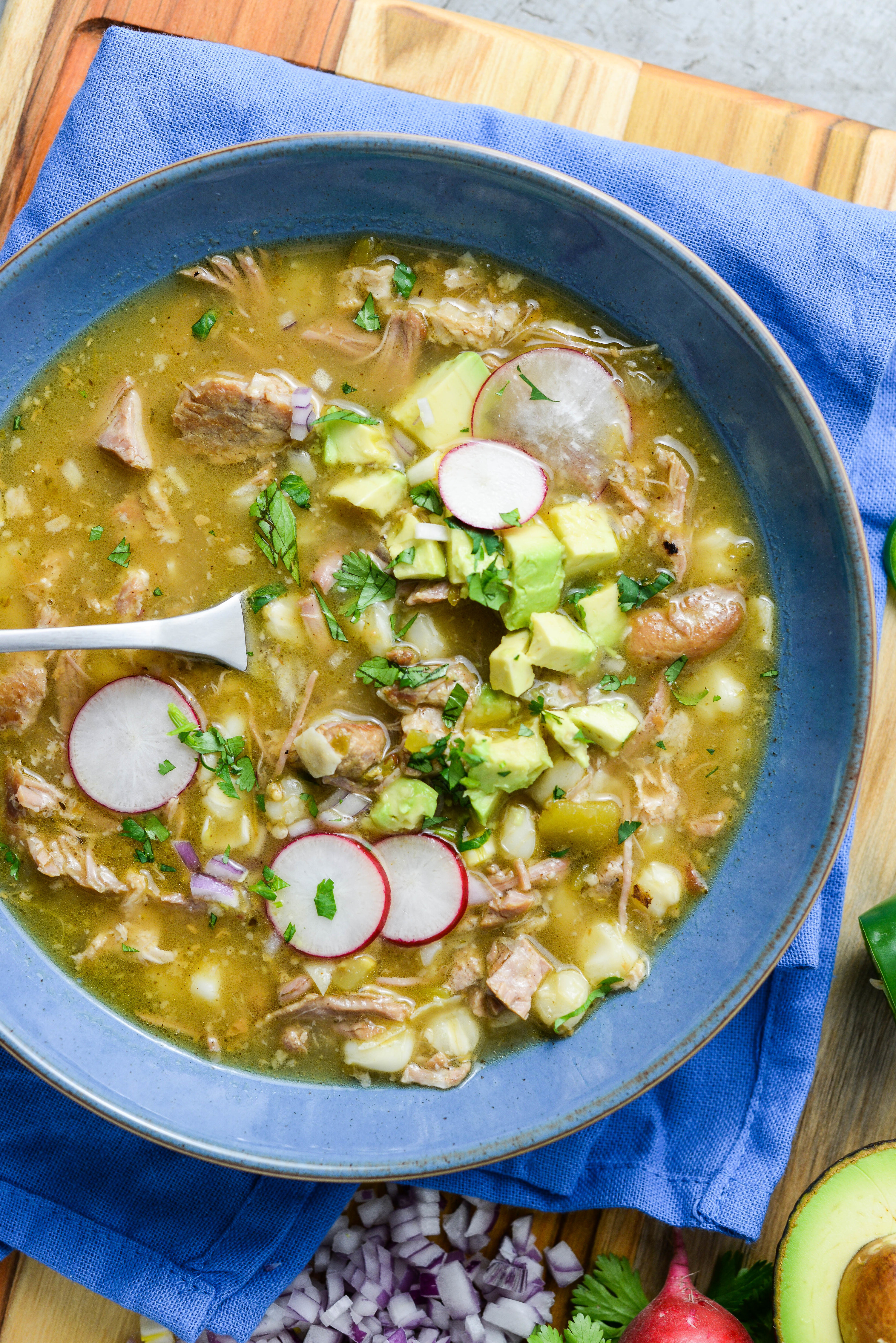 A bowl of green pozole with pork, hominy, radishes, chopped red onion, and avocado.