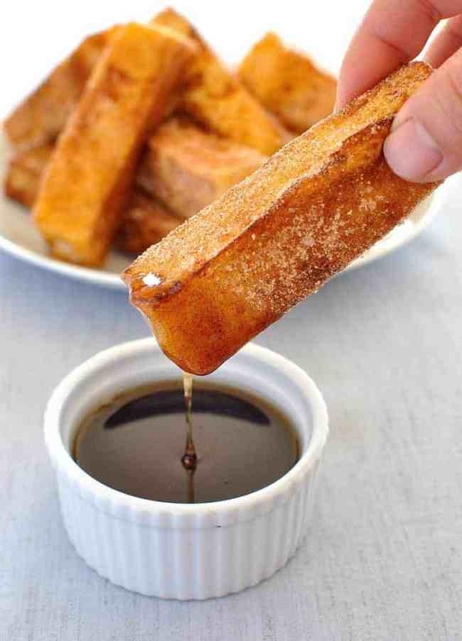 A french toast stick with cinnamon sugar being dipped into a ramekin of maple syrup.