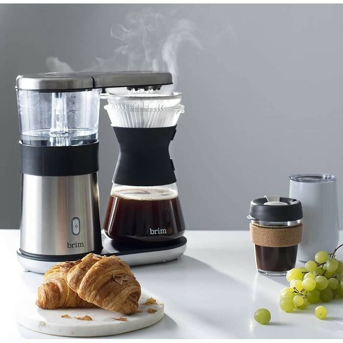 a silver pour-over coffee maker on a counter with croissants, grapes, and coffee in to-go cups