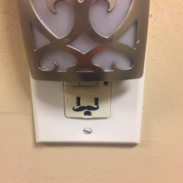 Reviewer's outlet with deodorizer plugged on top and mustache placed on bottom, making the outlet look like a face with a large hat on 