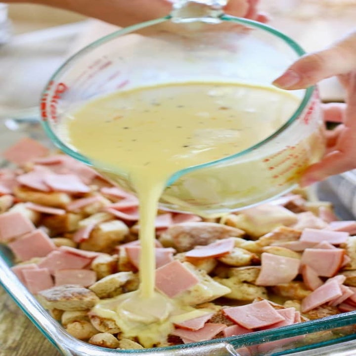 Pouring an egg mixture onto torn English muffins and Canadian bacon in a baking dish. 