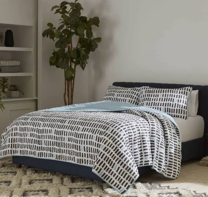 White quilt with gray and blue detailing on gray full size bed, white sheets underneath