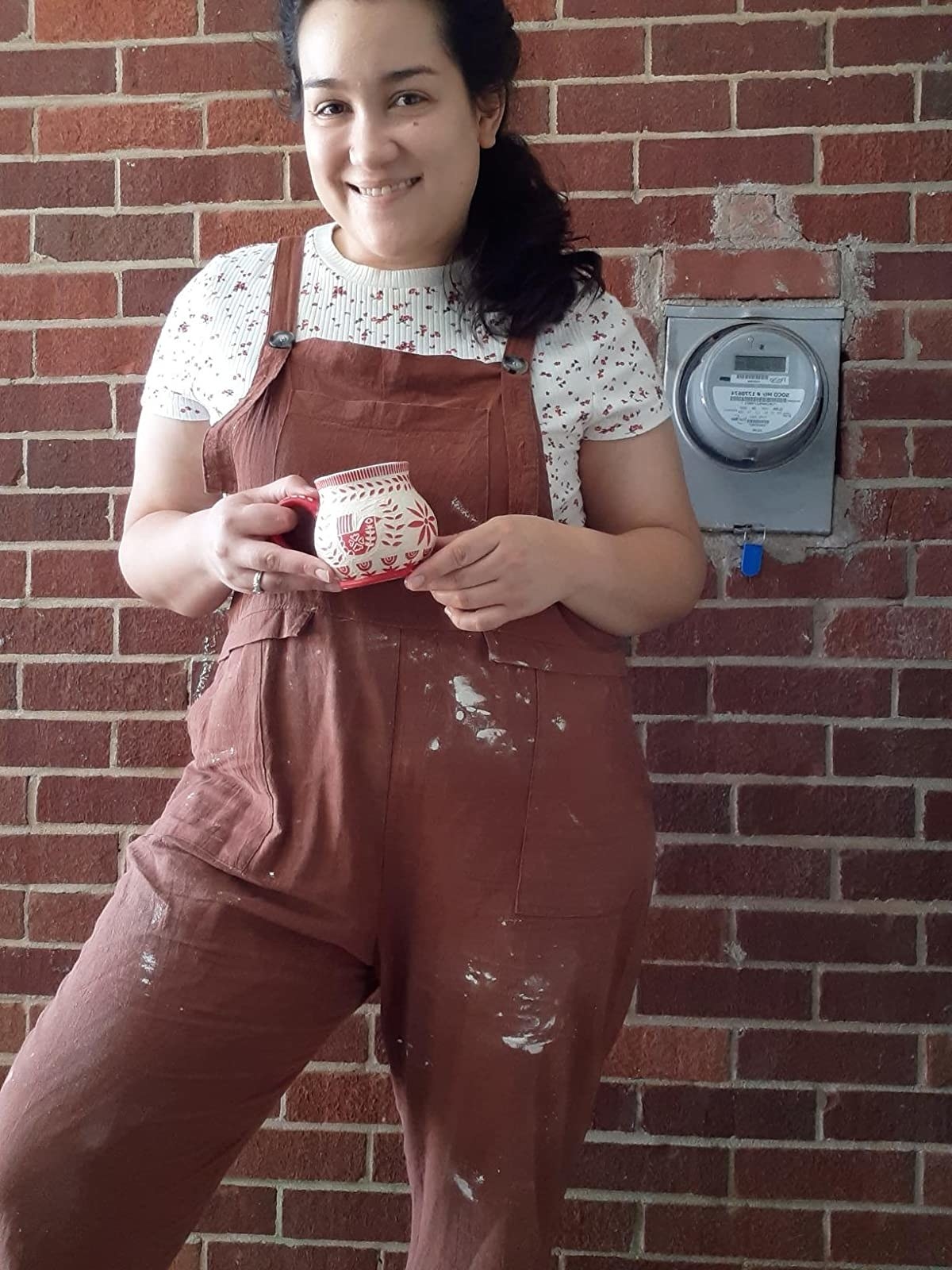 A reviewer wearing overalls splattered with clay spots layered over a T-shirt holding a small ceramic cup 