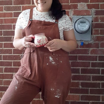 A reviewer wearing overalls splattered with clay spots layered over a T-shirt holding a small ceramic cup 