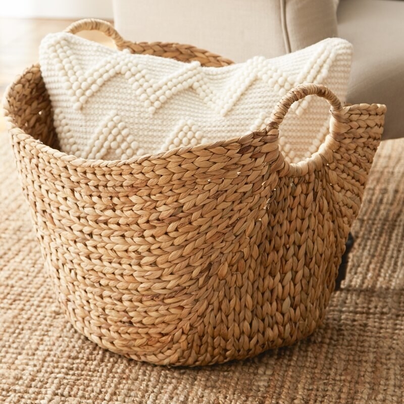a tan braided wicker basked with handles holding a pillow