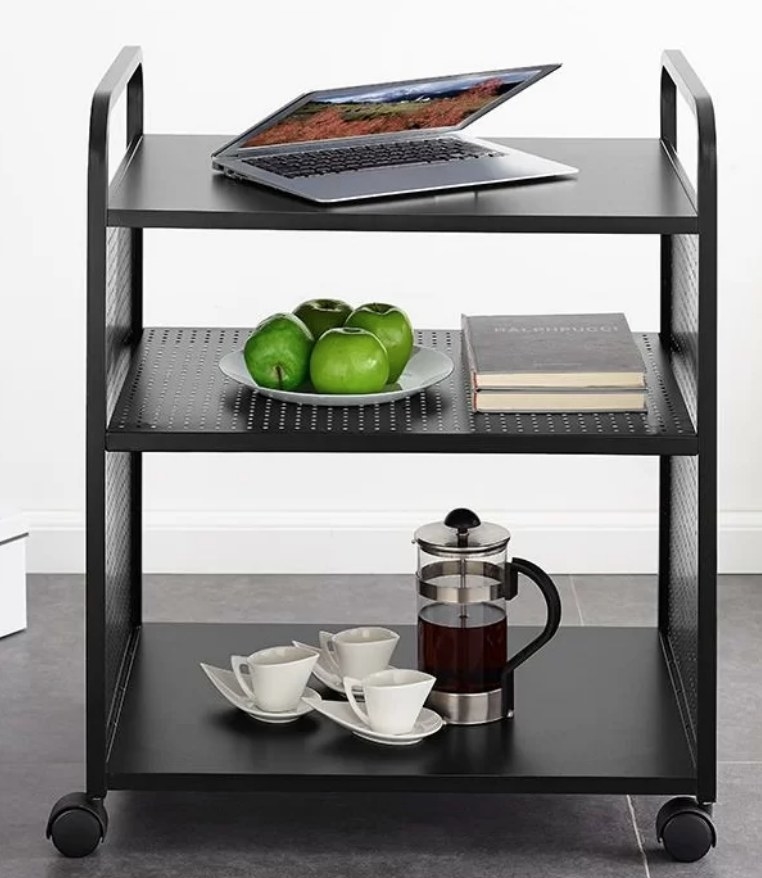 Black three tiered bar car with wheels, laptop on top shelf, apples and books on second shelf, french press and three coffee mugs on bottom shelf