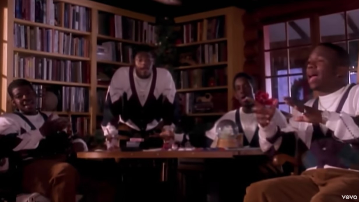 Boyz II Men wear matching Christmas cardigans and sing while sitting around a table in front of a bookshelf