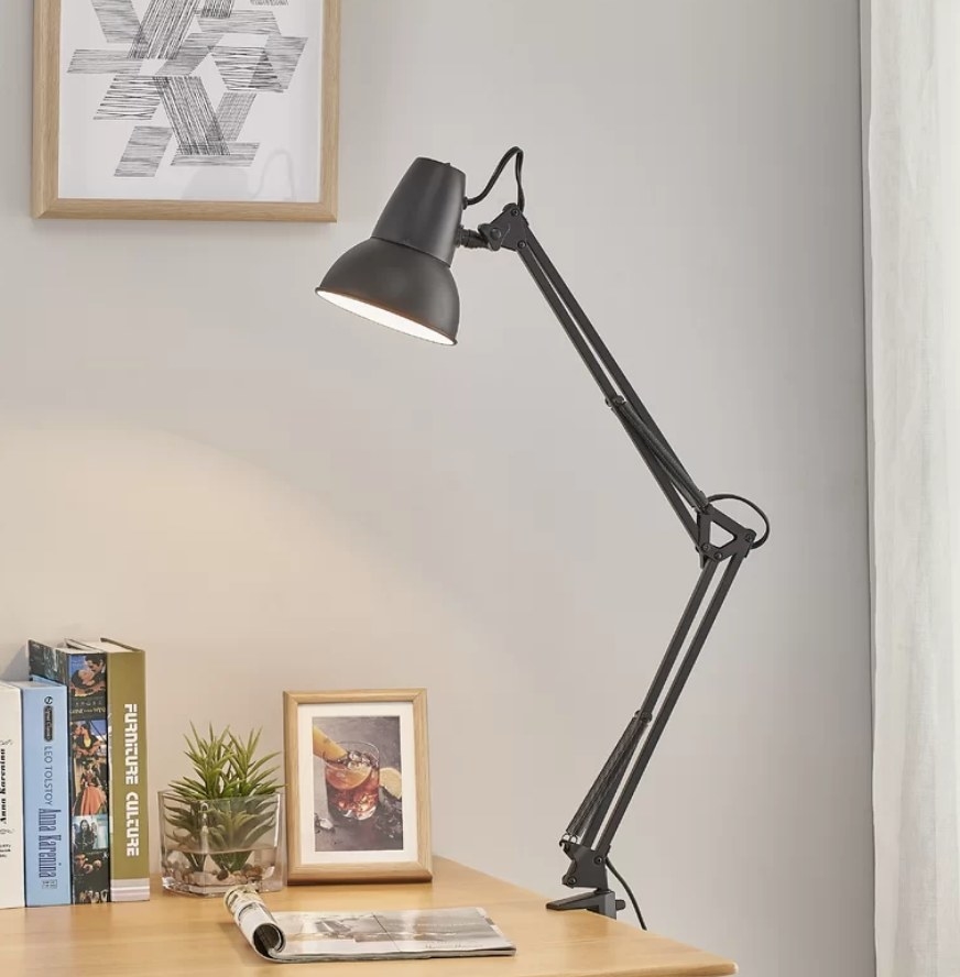 Black lamp clamped on edge of wooden desk that has a picture frame, open magazine, plant and books 