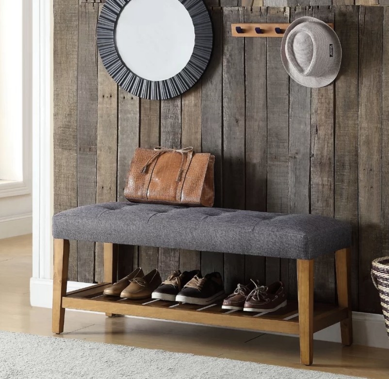 Gray cushioned bench seat with wooden frame and shelf that has three pairs of shoes underneath. 