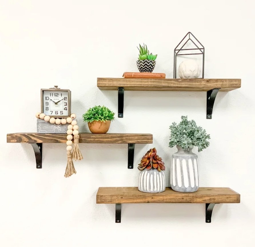 Three wooden floating shelves with black hinges, various knick knacks and planters on each shelf