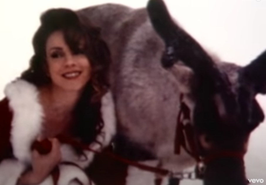 Mariah Carey wears a red outfit with fluffy white trim and bends down next to a reindeer