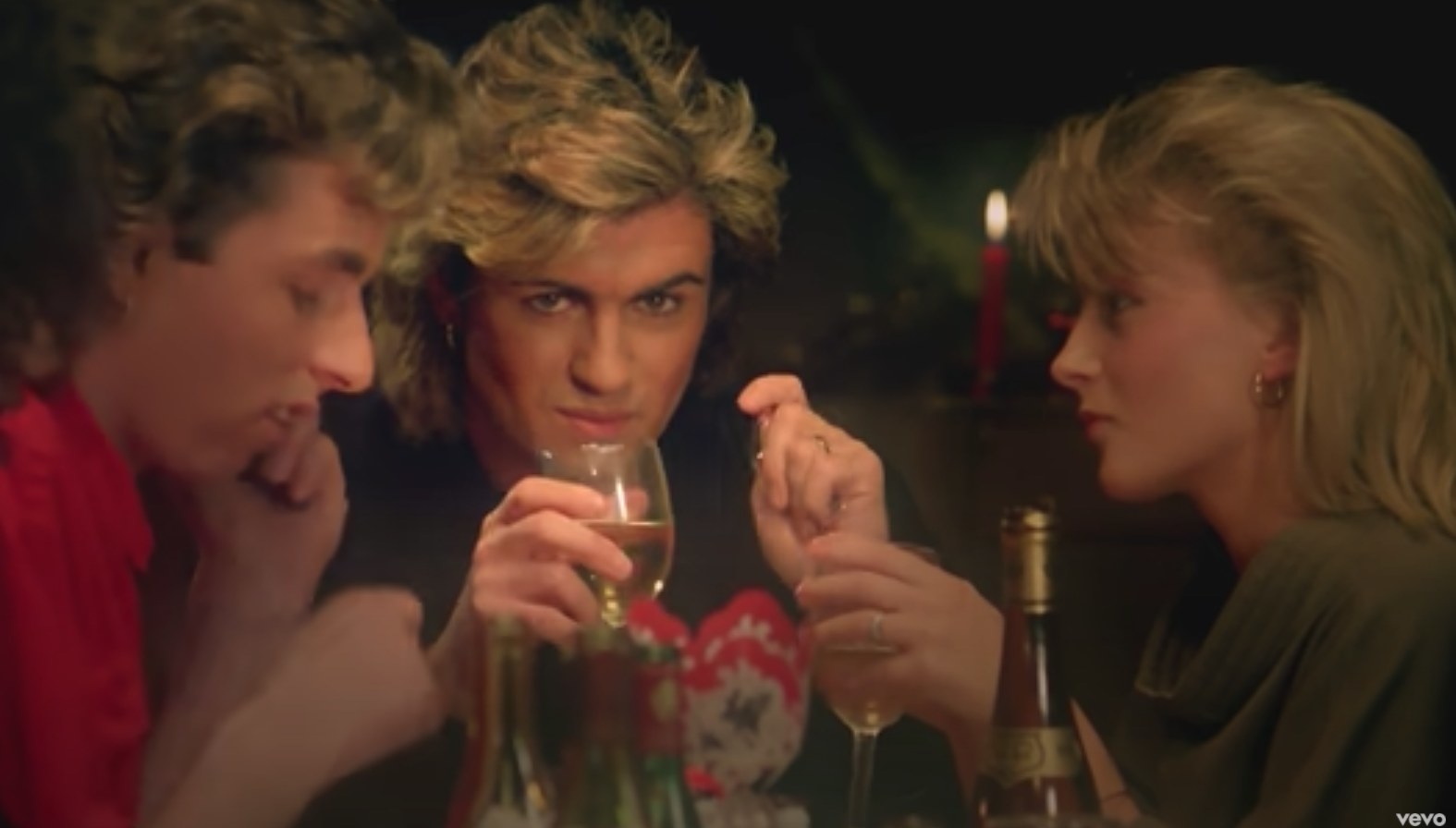 George Michael stares intently at someone over a wine glass; in front of him a man and a woman carry on a conversation
