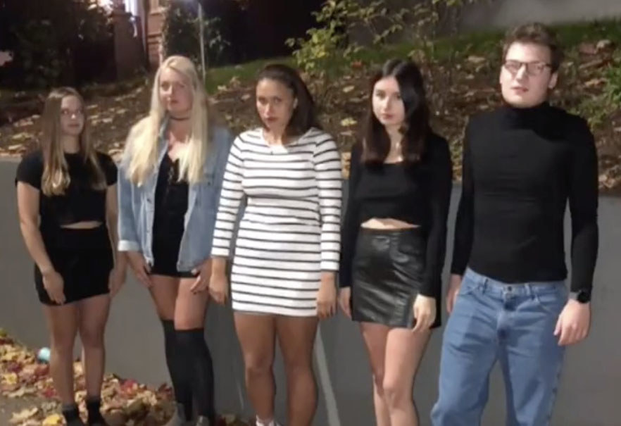 A group of friends dressed in black emo clothing stand in front of a wall