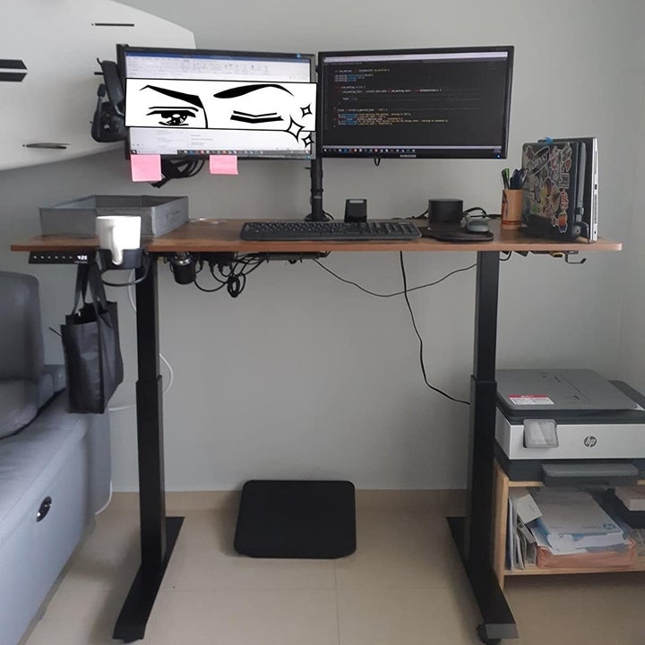 The brown and black standing desk supporting two monitors, a keyboard, and a collection of office supplies 