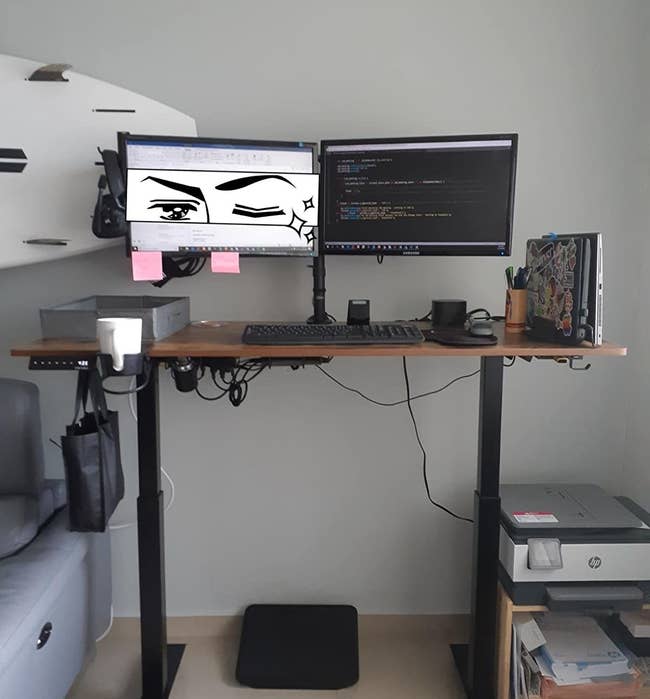 The brown and black standing desk supporting two monitors, a keyboard, and a collection of office supplies 