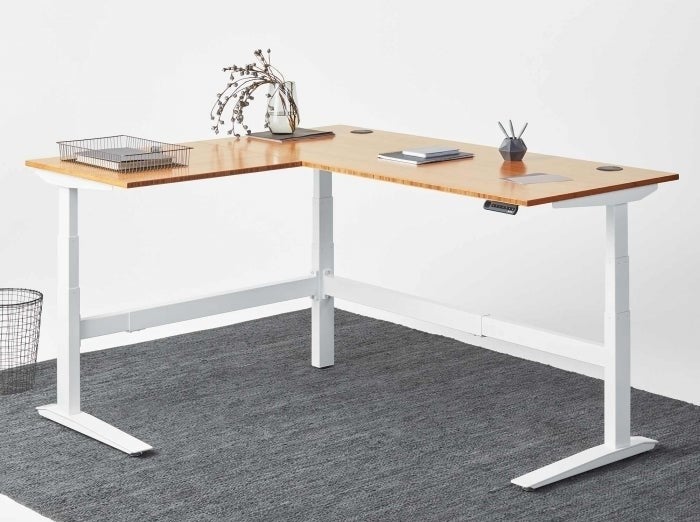 An L-shaped desk with a birchwood top, white legs, and a small control panel for adjusting the height 