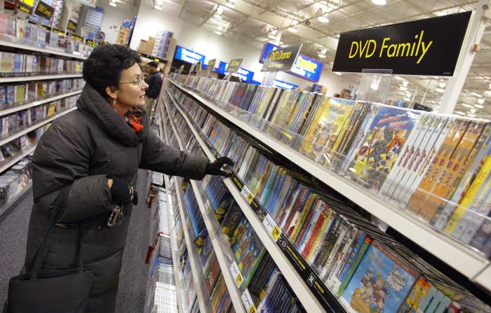 A photo of an older woman in a black winter coat looking DVDs in the DVD Family section of a Best Buy store