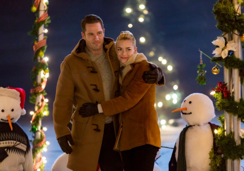 Still from The Mistletoe Promise: Jaime King and Luke McFarlane embrace surrounded by snowmen and Christmas decorations