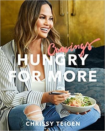 Chrissy Teigen sitting and eating a bowl of noodles on the front of her cookbook