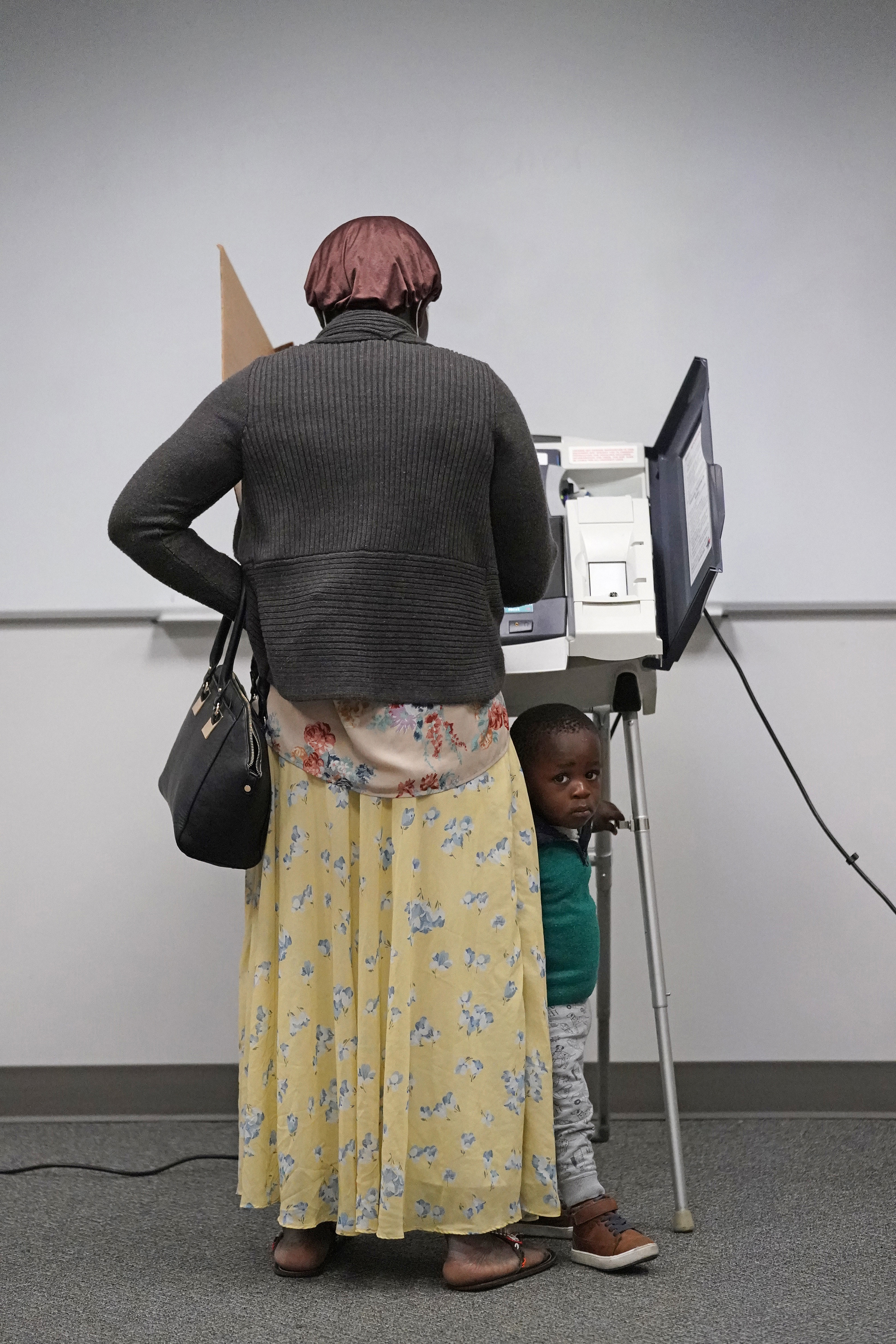 Woman with her back to the camera voting as her son peeks around her legs. 