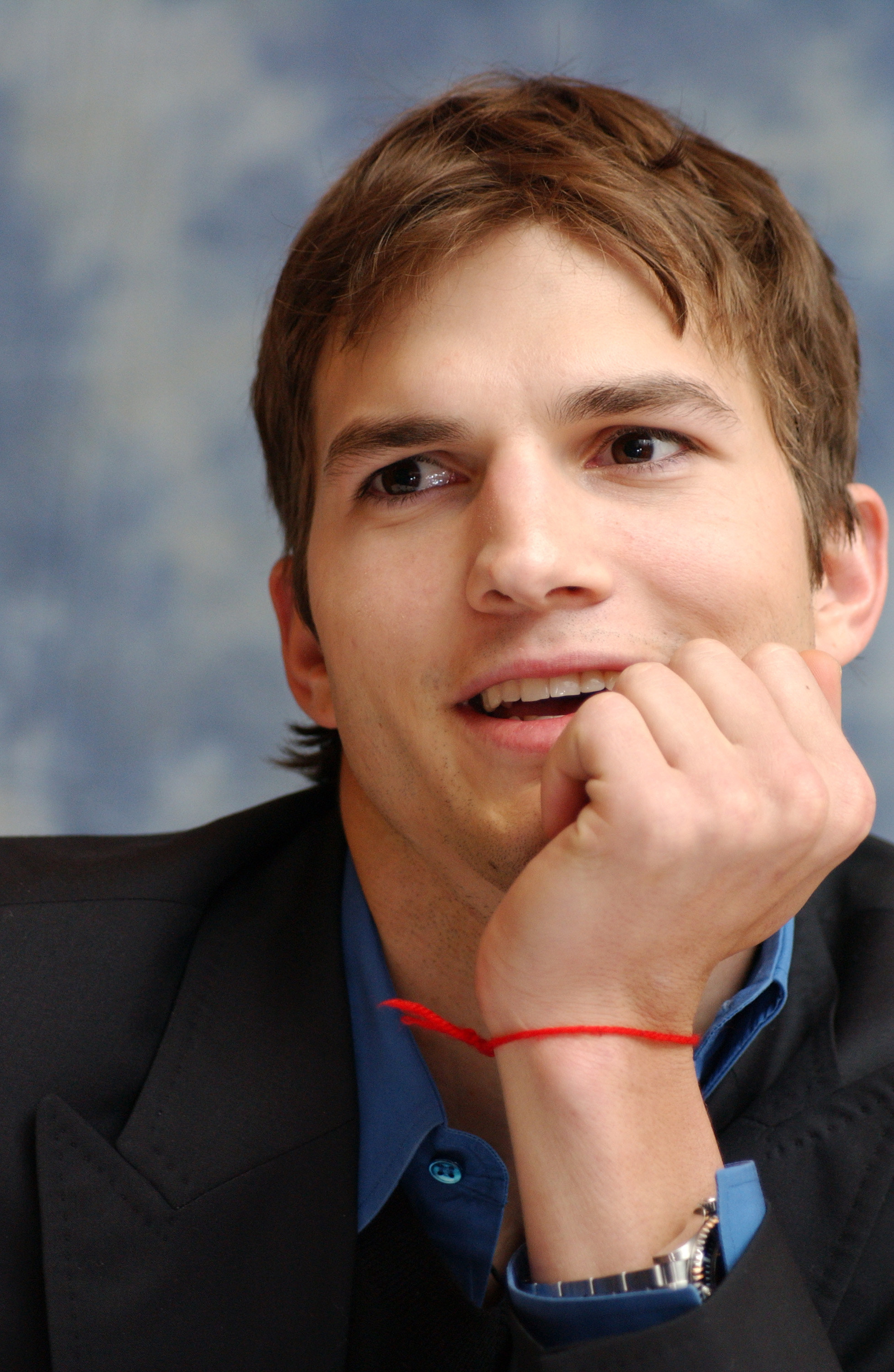 Ashton Kutcher at a press conference with a his head on his hand and a red string around his wrist 