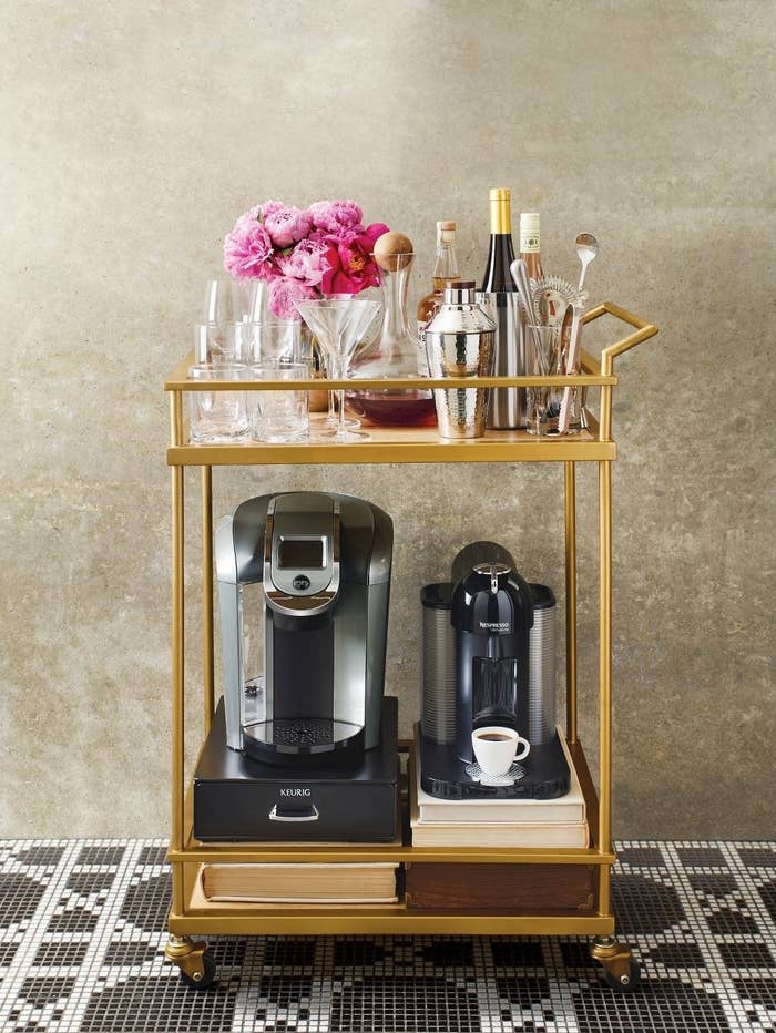 Wood and gold bar cart with black wheels carrying coffee machines on the bottom and cocktail shakers on top