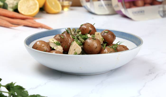 A bowl of whole little potatoes covered with chicken, green beans and green herbs. There are other bright, colorful vegetables surrounding the plate of food.