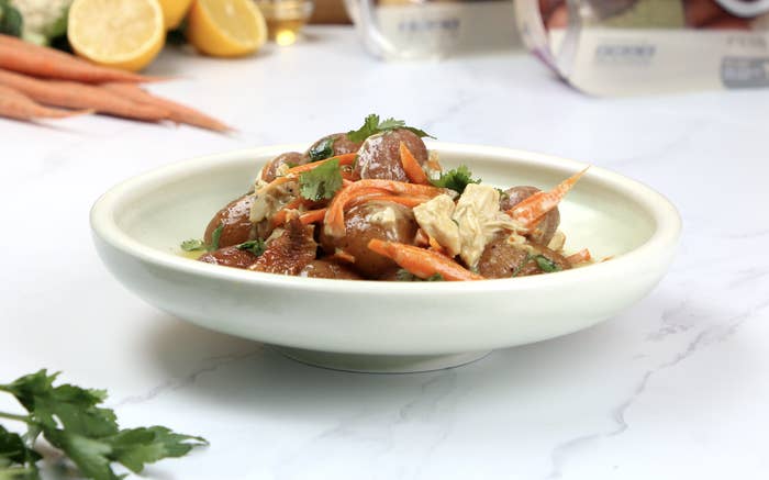 A bowl of whole little potatoes with pieces of chicken, carrot and cilantro on top. There are other bright, colorful vegetables surrounding the plate of food.
