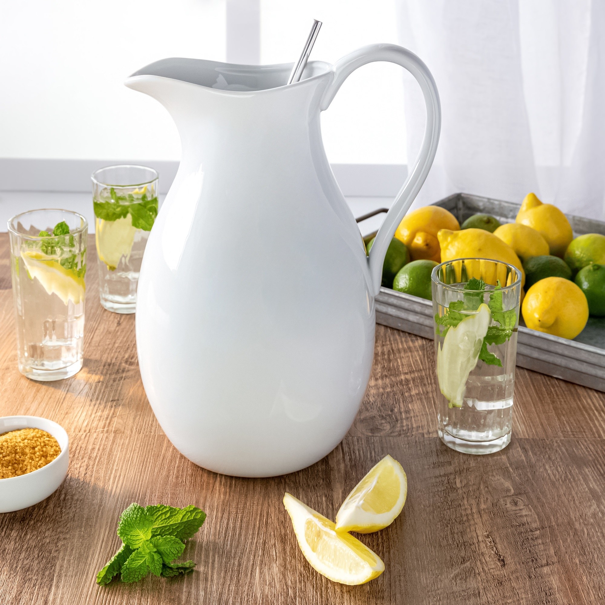 A white pitcher on a table, surrounded by lemons, limes, and mint