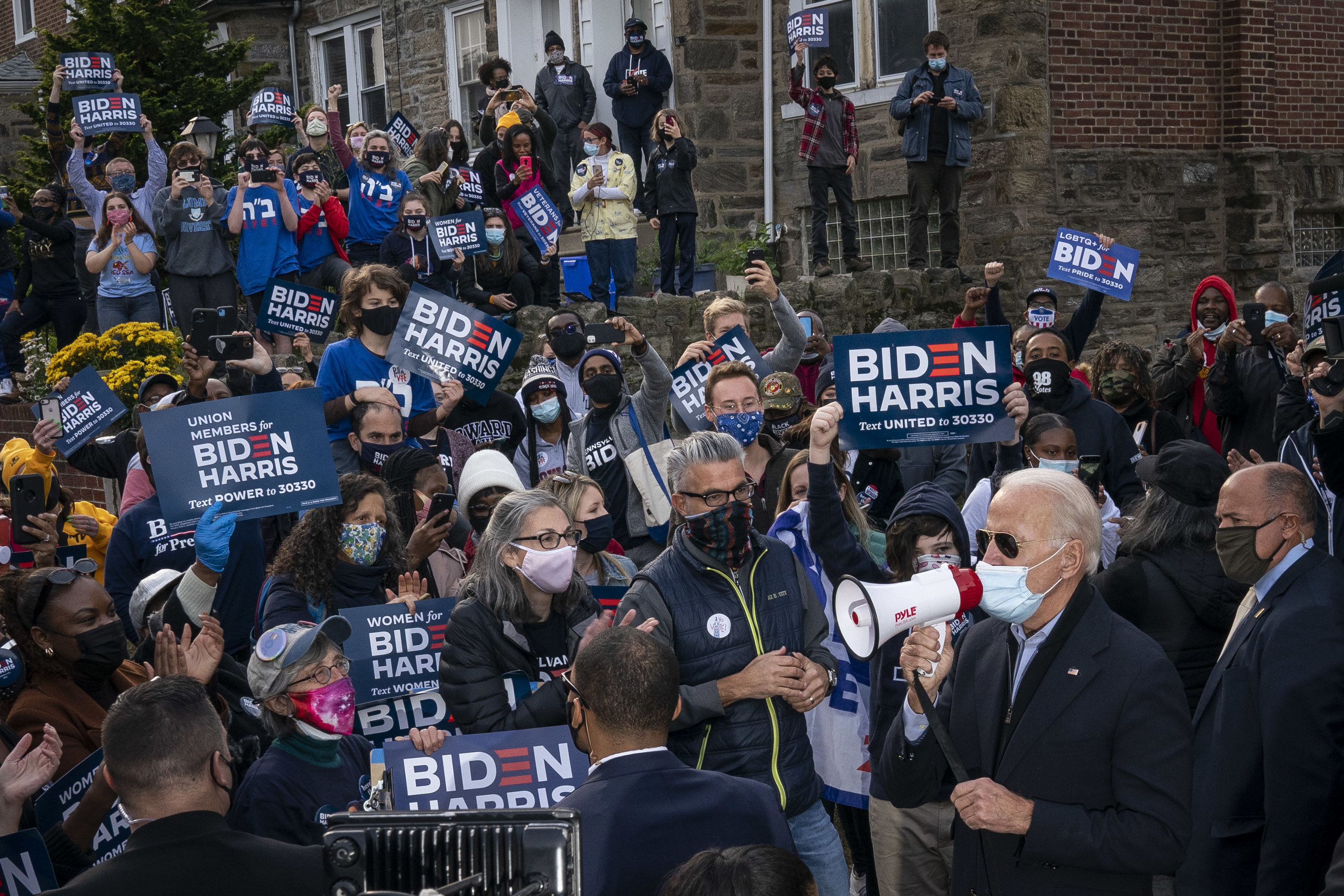 Joe Biden discursando em um megafone em frente à uma multidão de apoiadores.