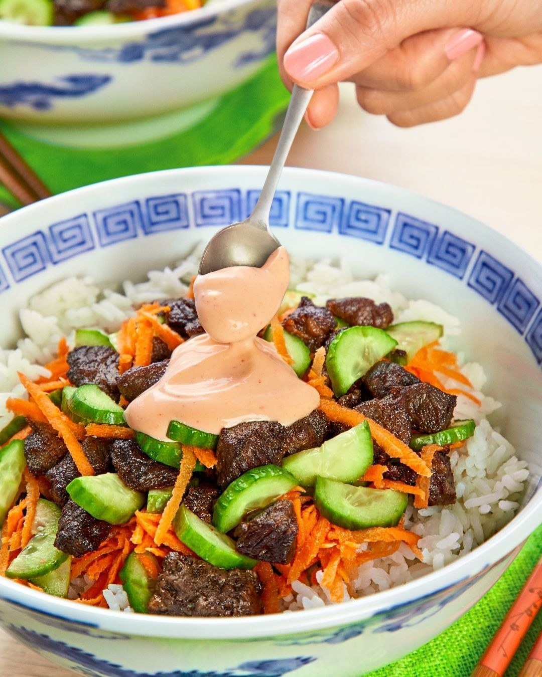 A hand drizzling sauce with a spoon onto a rice bowl topped with meat and vegetables