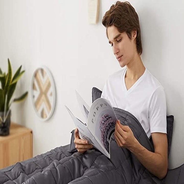 Model sitting in bed under a weighted blanket reading a magazine 