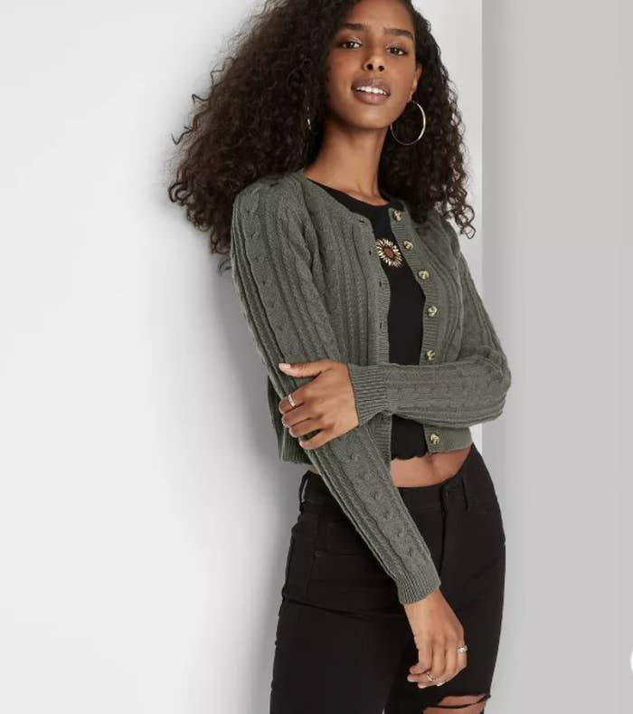 A model is wearing an olive green, cropped cardigan with buttons that is opened.