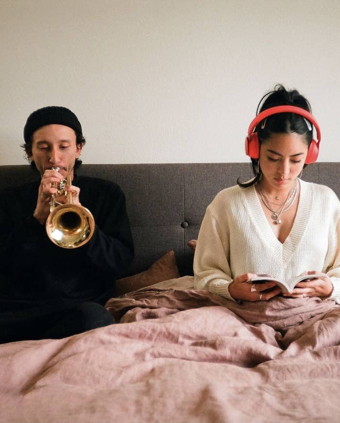 Person is sitting in bed with headphones in while the other person plays the trumpet next to them