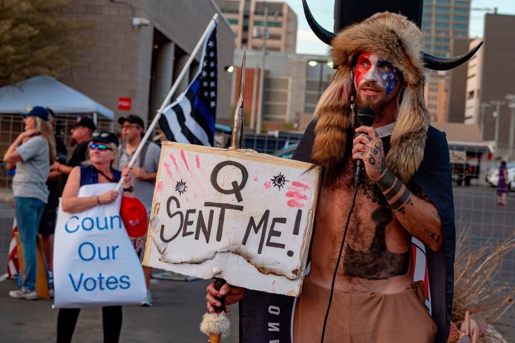 Woman waving a &quot;Blue Lives Matter&quot; flag stands near a wearing wearing a fur hat with horns speaks into a mic while holding a sign that says &quot;Q sent me&quot;