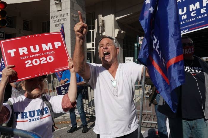 Trump supporter yelling as a woman holding a sign that says, &quot;Trump, Keep American Great! 2020&quot; stands next to him