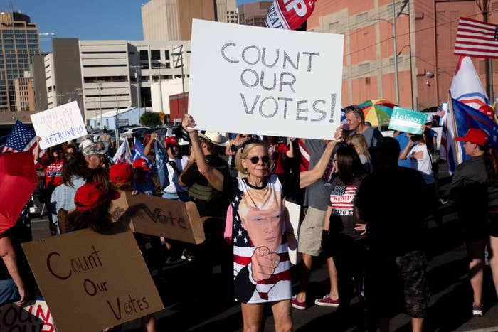 Woman wearing t-shirt dress with image of Donald Trump&#x27;s face holds sign saying, &quot;Count our votes!&quot;